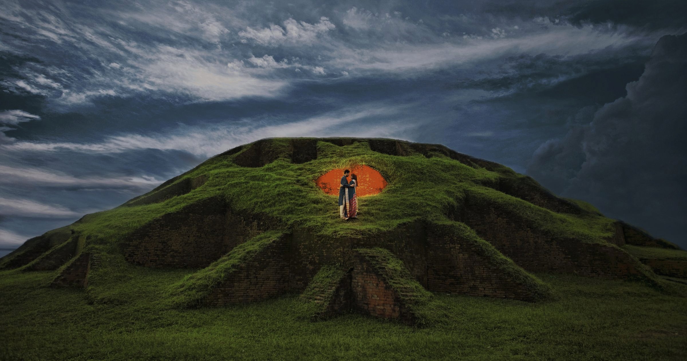 Bride and groom embracing inside a glowing brick archway atop a grass-covered hill at dusk